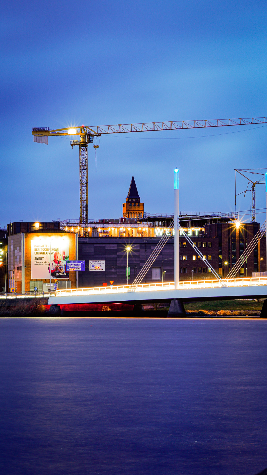 Construction cranes and a lit bridge over water in a Swedish city at dusk